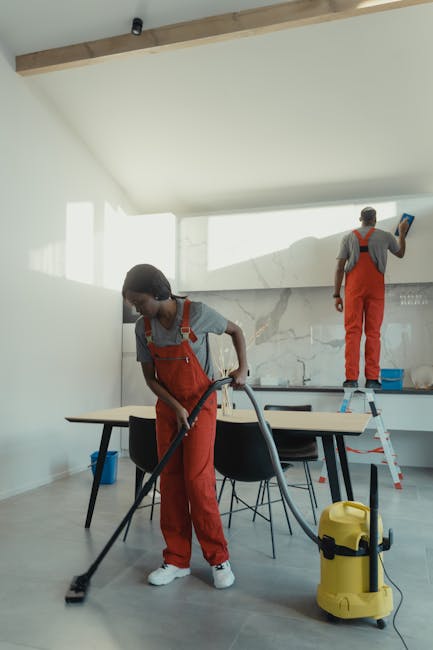 Two professional cleaners from West Kensington Cleaners are performing surface cleaning and deep cleaning tasks in a modern kitchen. The female cleaner in the foreground is vacuuming the floor with a yellow vacuum cleaner, wearing a grey t-shirt and red work overalls. In the background, a male cleaner is standing on a small step ladder, using a spray bottle to apply surface sanitisation or cleaning solution to the marble backsplash behind the countertop. The room features a white wall, a wooden beam on the ceiling, and a sleek design with a minimalist style, including a black dining table with chairs, and visible stainless steel appliances. Natural light fills the space, highlighting the cleanliness and well-maintained surfaces, emphasizing the service quality provided by West Kensington Cleaners for residential or commercial cleaning purposes.