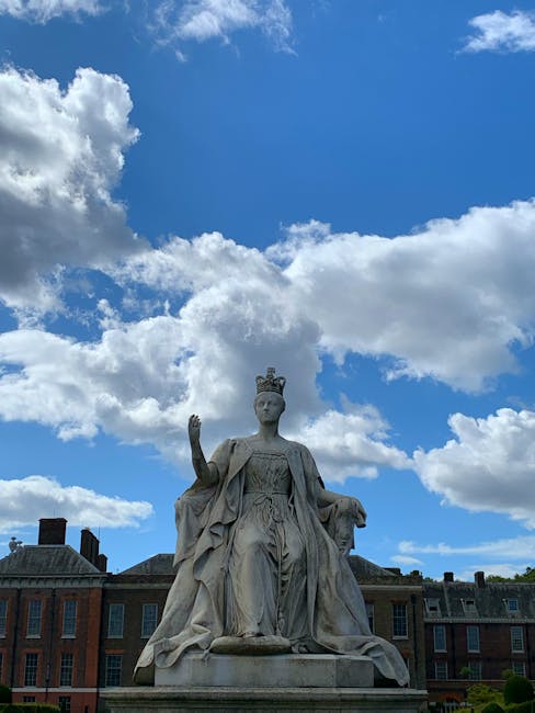 A historic stone statue of a regal woman seated outdoors, wearing a crown and flowing robes, situated on a base in front of a row of brick buildings with chimneys. The statue exhibits detailed carvings, with one hand raised as if in blessing and the other resting on her lap. The surrounding environment features a bright blue sky with scattered white clouds, and the area appears clean and well-maintained. The lighting highlights the intricate textures of the stone and the serene expression on the statue's face, capturing a scene of architectural heritage and outdoor artistry, emphasizing the importance of surface cleaning and preservation that West Kensington Cleaners offers in residential and commercial spaces.