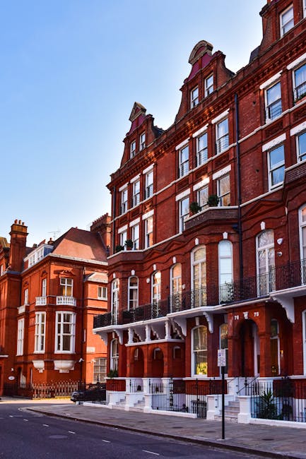 Image of a multi-story red brick residential building on a city street, featuring large windows with white framing, some with decorative arches, and black wrought iron balconies. The building has a white decorative cornice along its roofline, and the entrance area is supported by white columns with a set of steps leading up to it. The street appears clean and quiet, with a clear blue sky overhead. This building showcases classic Victorian architectural style, commonly found in West Kensington, and emphasizes the importance of maintaining external surfaces through regular surface cleaning and deep cleaning services, as offered by West Kensington Cleaners, to ensure hygiene and aesthetic appeal.