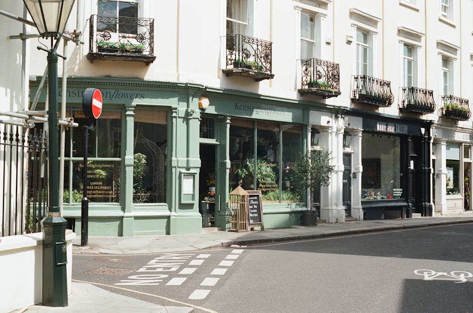 Exterior view of a charming, multi-story building on Lillie Road in West Kensington featuring a pastel green shopfront with large glass windows displaying a variety of plants and flowers, which are maintained in pots outside the store. The building has white walls and ornate black metal balconies on the upper floors, each with flower boxes. The sidewalk is clean and smooth, with a bicycle lane marked nearby. A black chalkboard sign stands just outside the shop, indicating information about the store's offerings. The scene is well-lit with natural daylight, emphasizing the neat and tidy appearance of the storefront, which reflects professional maintenance and cleaning standards. West Kensington Cleaners provides domestic cleaning and surface sanitisation services to maintain such pristine exteriors.