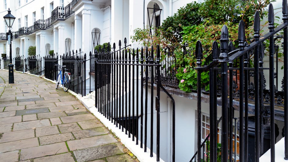 An exterior view of a row of white terraced houses on Lillie Road in West Kensington, with a paved sidewalk in the foreground. The sidewalk is composed of rectangular stone slabs, some showing signs of moss or weathering. Black wrought iron railings with pointed finials run alongside the houses, enclosing small front gardens with greenery, flowering plants, and climbing vines. A blue bicycle is parked against one of the railings. Soft natural lighting illuminates the scene, emphasizing the clean, well-maintained surfaces of the pavement, railings, and building facades. This image exemplifies meticulous surface cleaning and maintenance typical of deep cleaning and sanitisation practices that West Kensington Cleaners consistently employ for residential exteriors.