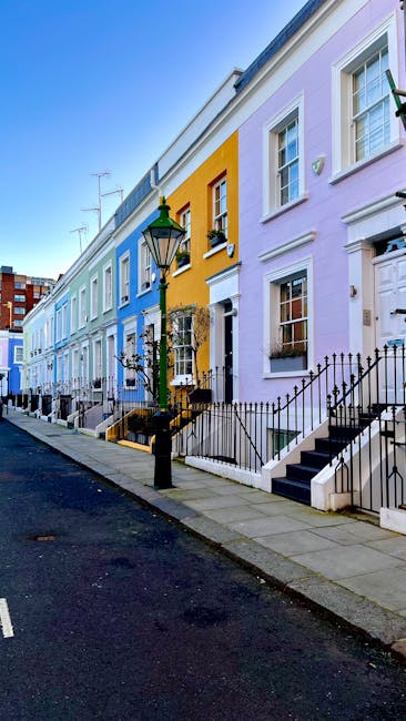 Exterior view of a charming, multi-story building on Lillie Road in West Kensington featuring a pastel green shopfront with large glass windows displaying a variety of plants and flowers, which are maintained in pots outside the store. The building has white walls and ornate black metal balconies on the upper floors, each with flower boxes. The sidewalk is clean and smooth, with a bicycle lane marked nearby. A black chalkboard sign stands just outside the shop, indicating information about the store's offerings. The scene is well-lit with natural daylight, emphasizing the neat and tidy appearance of the storefront, which reflects professional maintenance and cleaning standards. West Kensington Cleaners provides domestic cleaning and surface sanitisation services to maintain such pristine exteriors.