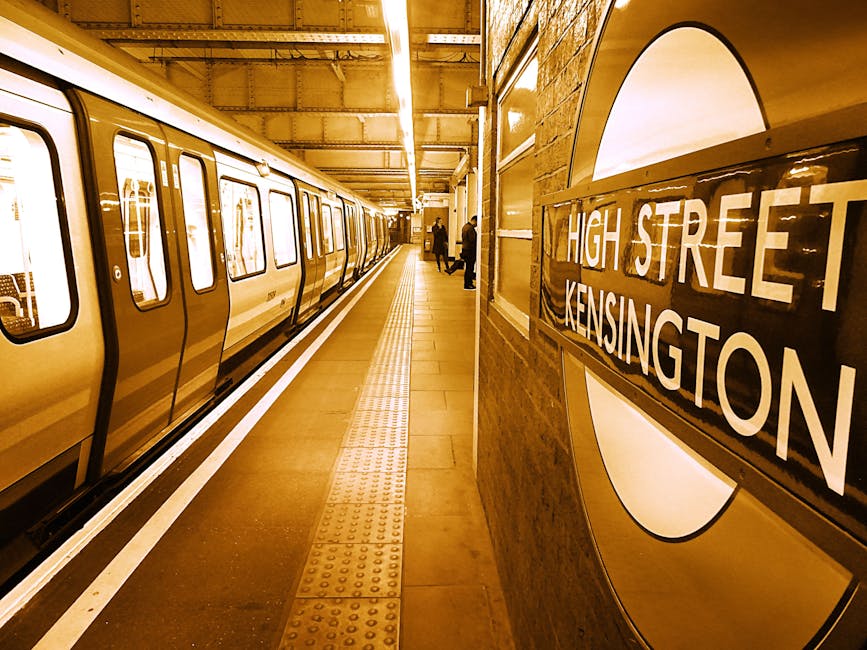 A view of the West Kensington underground station platform showing a train with closed doors on the left, with a yellowish-brown exterior, and a brick wall on the right featuring a large round sign with the station's name, 'High Street Kensington', in bold white letters against a black background. The platform surface is clean, with tactile paving strips near the edge for safety, illuminated by overhead lighting that creates a warm glow. Passengers are waiting at a distance, and the overall scene reflects a modern, well-maintained transit environment associated with domestic cleaning and maintenance standards emphasized by West Kensington Cleaners.
