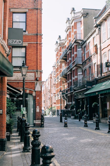 A quiet cobblestone street in West Kensington lined with red brick and stucco multi-story buildings. The buildings feature large windows and small balconies, with some storefronts displaying awnings and signage. The street is bordered by black bollards, with a vintage lamppost visible on the left side. Natural daylight illuminates the scene, highlighting the clean, well-maintained surfaces of the building exteriors. This setting reflects a typical area where residential and commercial cleaning services, such as those offered by West Kensington Cleaners, may be required to maintain hygiene and appearance in urban spaces.