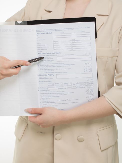 A person dressed in a beige coat is holding a clipboard with a form titled 'Home Insurance' on it, featuring sections for personal details, insurance history, and property information. The individual is using a black pen to point at the form, which is held in one hand, while the other hand supports the bottom of the clipboard. The scene is well-lit with a plain white background, highlighting the form and the person's attire. This image relates to surface cleaning or maintenance activities expressed through document review, associated with professional cleaning standards emphasized by West Kensington Cleaners as part of their licensing and insurance checklist for domestic cleaning in W14, West Kensington.