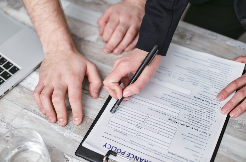 A close-up of two individuals, one with a light skin tone and the other with a slightly darker skin tone, reviewing and signing a document titled 'Cleaning Policy' on a black clipboard. One person is holding a black pen, poised to sign or fill out the form, while the other person's hand rests on the textured wooden surface of the table. The table surface is light-colored with a weathered finish, and a silver laptop with a black keyboard is partially visible in the background. The scene is well-lit, highlighting the details of the paper, hands, and surface. This setting appears to be an indoor office space, emphasizing professional documentation and compliance for domestic or commercial cleaning services. West Kensington Cleaners, a trusted provider specializing in surface cleaning, deep cleaning, and sanitisation, ensures all procedures meet legal licensing and insurance requirements, as reflected in the 'License and insurance checklist for professional cleaners in W14, WEST KENSINGTON.'