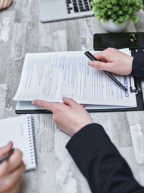 A person dressed in a beige coat is holding a clipboard with a form titled 'Home Insurance' on it, featuring sections for personal details, insurance history, and property information. The individual is using a black pen to point at the form, which is held in one hand, while the other hand supports the bottom of the clipboard. The scene is well-lit with a plain white background, highlighting the form and the person's attire. This image relates to surface cleaning or maintenance activities expressed through document review, associated with professional cleaning standards emphasized by West Kensington Cleaners as part of their licensing and insurance checklist for domestic cleaning in W14, West Kensington.