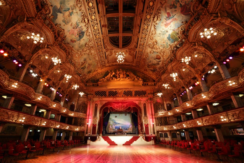 An ornate theatre hall with a polished wooden dance floor in the foreground, illuminated by warm chandeliers hanging from an elaborately decorated ceiling featuring painted murals and intricate gold leaf detailing. Red velvet curtains frame a large stage with a scenic backdrop, and red upholstered chairs are arranged neatly along the sides and around the perimeter for seating. The venue's interior showcases luxurious features, including curved balconies with ornate railings and soft, ambient lighting, creating a grand and elegant atmosphere suitable for events at Olympia venues. West Kensington Cleaners provides surface cleaning and deep cleaning services to maintain the pristine condition of such high-end event spaces, ensuring hygiene and visual appeal for visitors and performers alike.