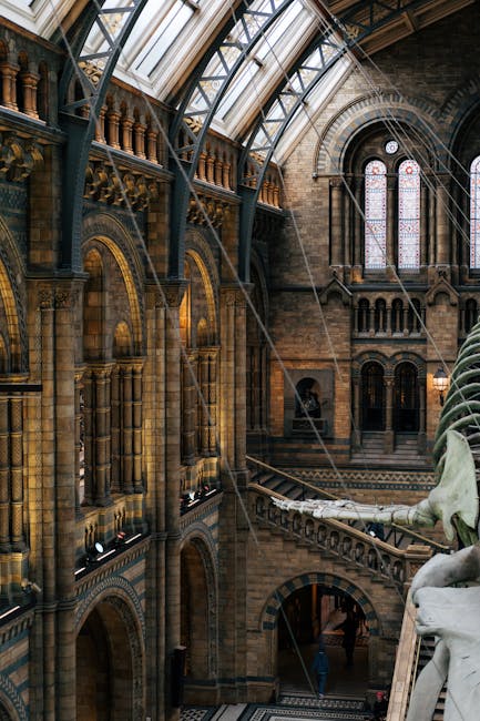 The interior of the Olympia venue showcases a historic architectural design with polished stone floors and intricate brickwork walls. Large arched stained glass windows allow natural light to fill the space, highlighting the detailed wood and metal railings along the mezzanine level. The high, vaulted glass ceiling, supported by metal framework, reflects a clean, dust-free surface, indicating recent surface cleaning and maintenance. The polished stone surfaces, including decorative staircases and columns, appear shiny and well-maintained. Visible cleaning tools, such as a cleaning spray and cloths, suggest ongoing surface cleaning processes. The overall space exudes a tidy and sanitised environment, with attention to hygiene typical of professional cleaning by West Kensington Cleaners, ensuring the venue remains pristine for events and visitors.