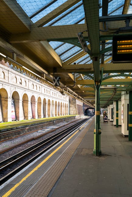 An indoor railway station platform at West Kensington station, showing clean, smooth concrete flooring with tactile yellow strips along the edge adjacent to the train tracks. The platform features Green metal support columns and a canopy with a glass and steel roof allowing natural light to illuminate the area. The station walls are lined with decorative brickwork and arched windows, some posters are visible on the walls, and a few passengers are waiting near the platform edge. The image emphasizes the pristine condition of the surfaces, reflecting thorough cleaning and maintenance by West Kensington Cleaners, which ensures a hygienic and safe environment for commuters. The area appears well-lit and free of dust or debris, highlighting the importance of surface cleaning and sanitisation in transportation spaces.