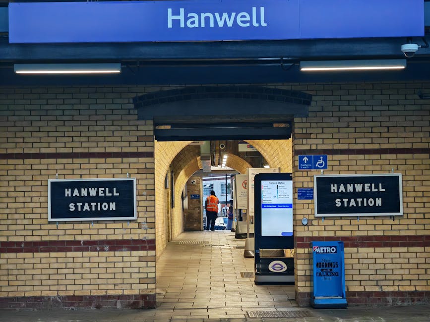 An indoor railway station platform at West Kensington station, showing clean, smooth concrete flooring with tactile yellow strips along the edge adjacent to the train tracks. The platform features Green metal support columns and a canopy with a glass and steel roof allowing natural light to illuminate the area. The station walls are lined with decorative brickwork and arched windows, some posters are visible on the walls, and a few passengers are waiting near the platform edge. The image emphasizes the pristine condition of the surfaces, reflecting thorough cleaning and maintenance by West Kensington Cleaners, which ensures a hygienic and safe environment for commuters. The area appears well-lit and free of dust or debris, highlighting the importance of surface cleaning and sanitisation in transportation spaces.