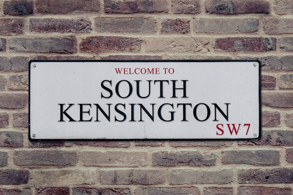 A rectangular metal sign affixed to a brick wall, featuring black and red lettering. The sign displays the text 'Welcome to South Kensington SW7,' with 'South Kensington' in large bold black font, 'Welcome to' in smaller red font above, and the postal code 'SW7' in red at the bottom right corner. The brick wall behind the sign is composed of uniformly textured reddish-brown bricks with visible mortar lines, providing a traditional and durable background. The sign and bricks are well-maintained, indicating regular cleaning and upkeep, typical in residential or commercial premises in West Kensington. West Kensington Cleaners emphasizes surface cleaning and sanitisation, ensuring surfaces like brick, metal, and signage remain clean and presentable for tenants and visitors.