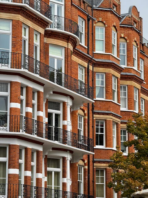 A close-up view of the exterior of a multi-story residential building showcasing red brick walls with white decorative horizontal bands and ornate architectural details. The building features large, white-framed sash windows with clear glass panes, some of which are partially open. The balconies have black wrought-iron railings, and the ground level displays a clean, well-maintained facade. Soft natural light highlights the texture of the brickwork and the shine of the windows, with a small tree with green and yellow leaves visible in the lower right corner. This setting exemplifies the typical architecture found in North End Road, West Kensington, emphasizing the importance of regular exterior surface cleaning to maintain both aesthetic appeal and building hygiene, as advocated by West Kensington Cleaners for domestic and commercial cleaning projects.